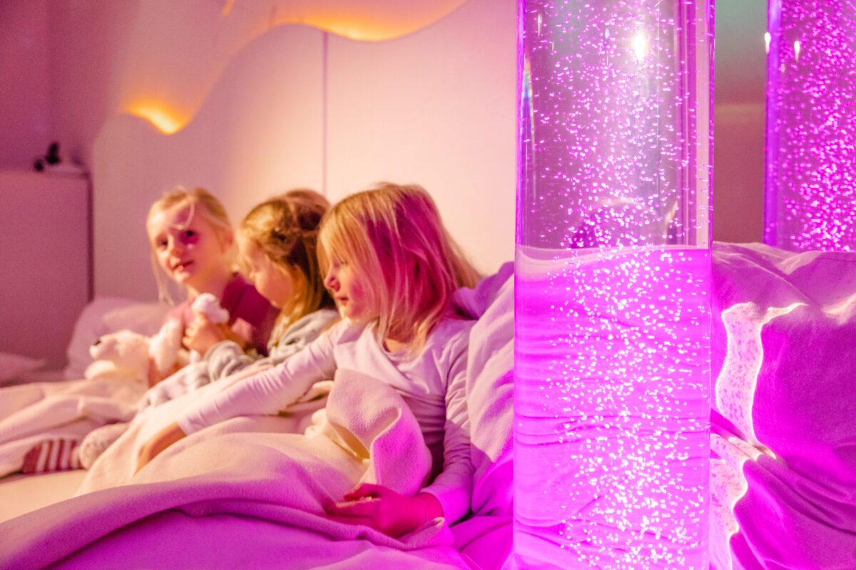 Spaß in der Kindertagesstätte Drei Mädchen sitzen in einem Bett, umgeben von Decken; sie kuscheln mit Plüschtieren und schauen auf einen beleuchteten Wasserzylinder, der pinke Blasen zeigt.