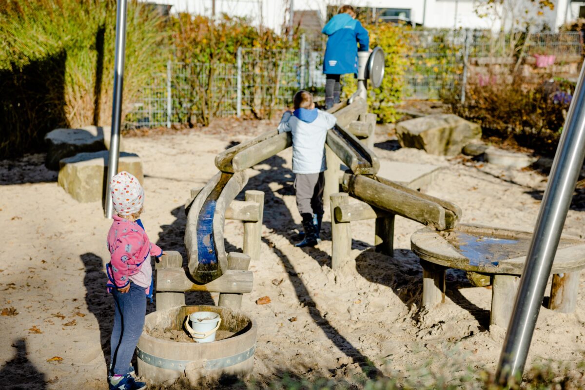 Spielplatz in der Kita Landwehr Zwei Kinder spielen in einem Sandspielbereich mit einer Holzspielkombination; ein Kind erkundet eine Wasserbahn, während das andere auf einem Kletterelement steht.