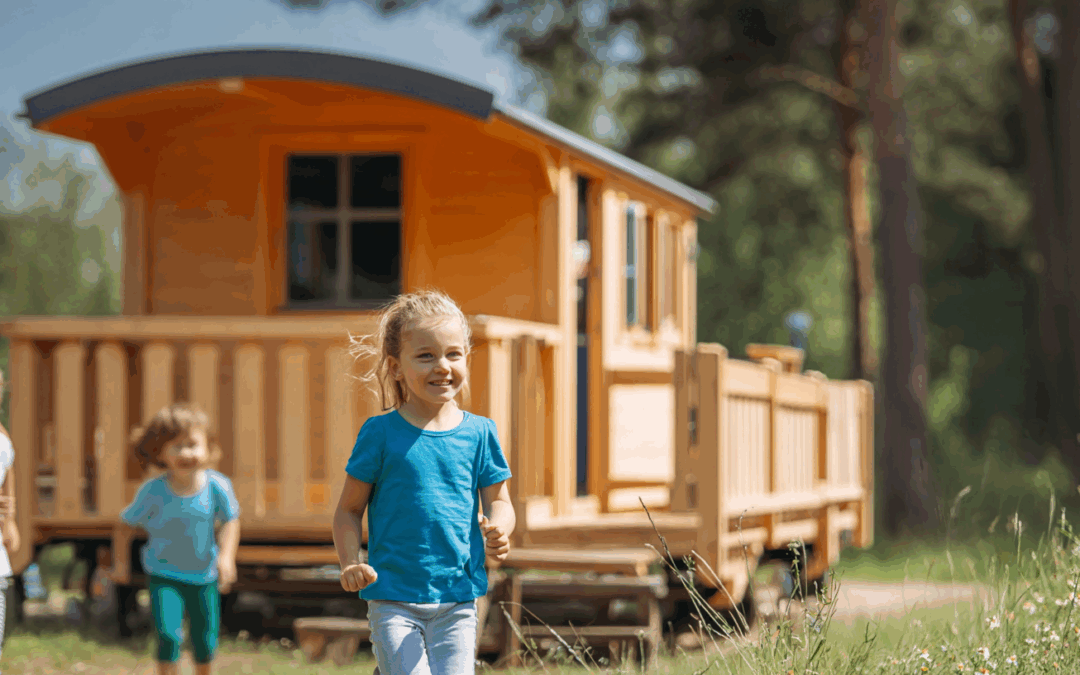 Ein fröhliches Mädchen in einem blauen T-Shirt läuft vor einem Bauwagen im Außengelände. Hinter ihm sind weitere Kinder zu sehen, die ebenfalls spielen.